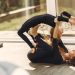Side view of cheerful mom in sports clothes lying on floor and teaching smiling daughter doing back bend in apartment in sunlight