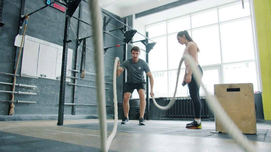 Energetic sportsman is battling ropes working out during sports training with coach young woman in modern gym. Active youth and workout concept.